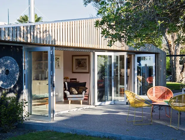 Single-storey contemporary dwelling or extension viewed from the garden, with a low-pitched roof, vertically slatted timber upper facade, dark painted exterior walls, and extensive glazed folding or sliding doors opening onto a paved outdoor sitting area. A furnished living room is visible through the glazing, while colourful outdoor wire-frame chairs and a small table occupy the terrace. Mature trees and planting surround the structure.