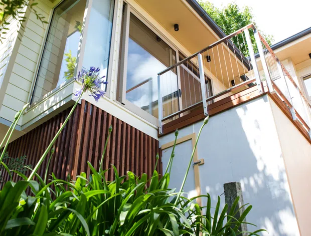Upward-looking exterior view of a multi-level residential building surrounded by dense garden planting. The facade combines pale horizontal weatherboard cladding, dark timber vertical screening, large glazed doors and windows, and a timber balcony with simple metal balustrading. A projecting roof eave with recessed downlights and dark roof edging is prominent.