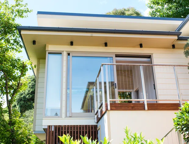 Exterior view of a contemporary single-storey home extension or facade, featuring cream horizontal cladding, a broad low-pitched roof overhang with recessed downlights, large glazed sliding doors, and a small balcony or deck with slim metal balustrades and timber detailing. The building is surrounded by lush vegetation.