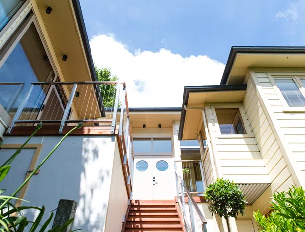 Upward-looking exterior view of a multi-level residential building with cream weatherboard cladding, balconies, extensive glazing, and an external stair leading to a glazed entry or upper level. The composition shows a bright contemporary coastal-style facade and courtyard-like access space.