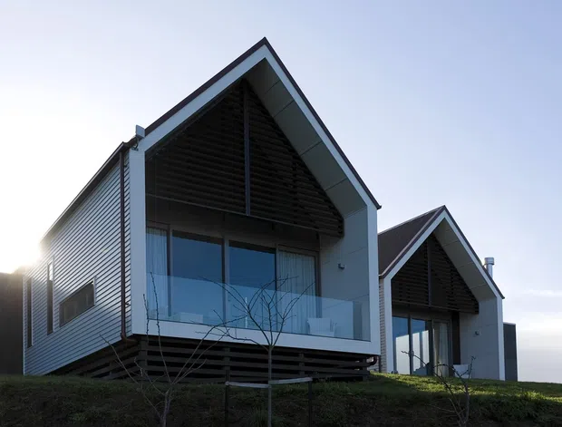 Exterior view of two compact contemporary gable-roofed dwellings on a gently sloping site. The buildings feature light horizontal cladding, dark timber-like soffits and louvred gable screens, generous glazed openings, and projecting upper-level balconies with clear glass balustrades.