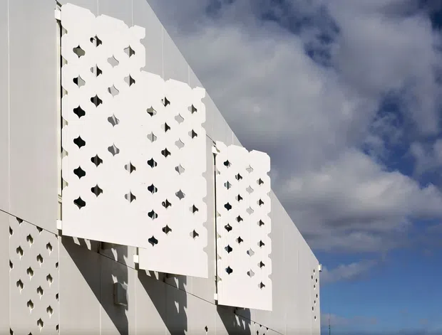 Exterior architectural detail of a contemporary white building facade featuring suspended decorative perforated screens with repeating geometric cut-outs. The screens cast sharp shadows across smooth light-coloured wall panels, with blue sky and clouds visible behind the structure.