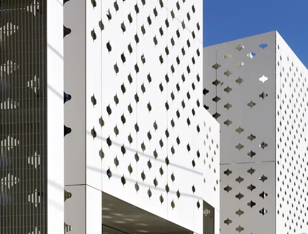 Exterior architectural photograph of a modern white multi-storey building composed of sharply folded and projecting volumes. The façades feature dense repeated decorative perforations in organic leaf- and star-like shapes, with a tall screened glazed or louvred section at the left. Strong sunlight creates deep shadows across the white surfaces against a clear blue sky.
