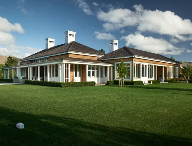 A large single-storey residential house viewed from the landscaped front lawn. The home has a low-pitched hipped roof with multiple white chimneys, extensive white-framed windows and glazed verandah-like galleries, light cream walls, and dark timber-toned roof shingles. It sits in a broad manicured lawn with distant hills visible behind it.