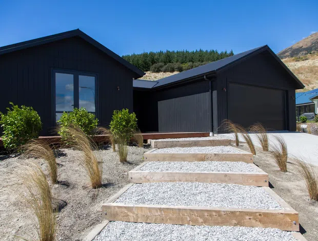 Exterior view of a contemporary single-storey dark-clad home with two steep gabled volumes, integrated double garage, and a landscaped gravel forecourt. Concrete stepping slabs lead through ornamental grasses toward the entrance and garage. The property sits against a dry, sloped hillside with trees in the background.