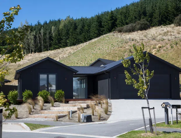 A contemporary single-storey dark-clad house set into a sloping, landscaped site. The building has multiple gabled roof forms, large glazed openings, and an integrated dark garage, with a pale concrete driveway and ornamental grasses in the foreground. Dense woodland and a steep grassy hillside form the backdrop.
