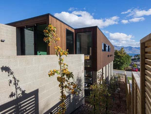 Exterior view of a contemporary multi-level residence with dark timber-clad upper volumes, exposed concrete block walls, and large glazed openings. A timber slat screen and landscaped narrow courtyard frame the foreground, while mountain scenery is visible beyond the property.