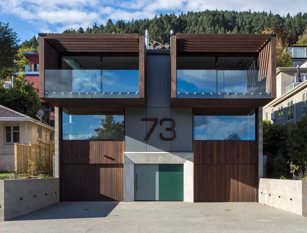 Modern multi-level residential building facade with two symmetrical upper balconies enclosed by clear glass balustrades, deep timber-clad roof projections, dark timber garage or entry doors, and a central light-grey concrete entry volume marked with the number 73. The building sits on a sloped suburban site with neighbouring homes and a wooded hillside behind it.