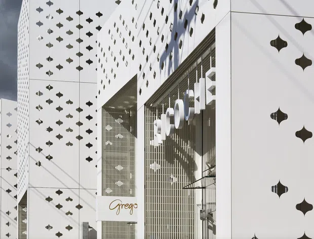 Exterior architectural photograph of a contemporary commercial building clad in white panels perforated with repeated quatrefoil-like openings. A tall glazed storefront and projecting entrance volume feature vertical metal screens, signage, and strong geometric shadows. The facade has a crisp, graphic appearance under a dramatic cloudy sky.
