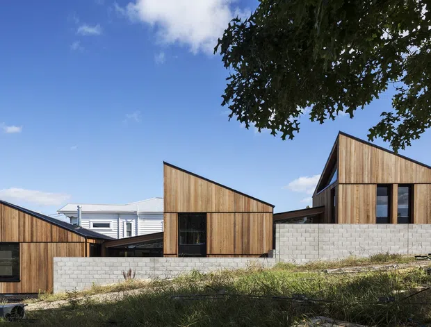 Exterior view of a contemporary multi-volume residential building on a sloped site. The composition features angular gabled roof forms, vertical timber-clad walls, dark-framed glazing, and pale masonry block retaining walls, with mature trees and a blue sky in the background.