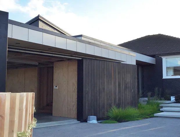 Exterior view of a contemporary residential frontage with a flat-roofed entrance or carport structure, dark vertical timber cladding, pale metal fascia panels, and a recessed timber door. A pitched roof is visible behind the modern addition, with concrete steps and low landscaped planting leading to the entry.
