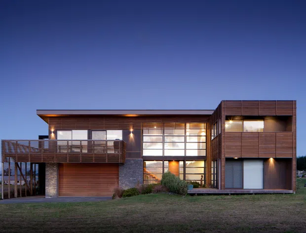 A contemporary multi-level residential house photographed at dusk, with a rectilinear flat-roofed composition. The facade combines dark horizontal timber cladding, extensive floor-to-ceiling glazing, stone veneer, and a projecting timber balcony with open horizontal screening. A garage and entry-level glazing are visible, with warm interior and exterior lighting contrasting against the blue evening sky.