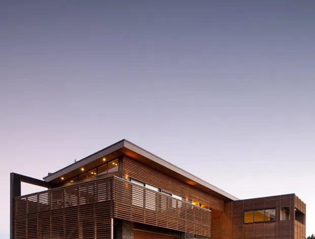 A contemporary multi-storey residential house photographed from the driveway at dusk. The building has a strong angular roofline, extensive horizontal timber screening and cladding, dark-framed glazing, a timber garage door, and a stone-clad section around the entry and garage. Low ornamental landscaping surrounds the paved driveway, while warm interior and exterior lighting creates a welcoming evening appearance.
