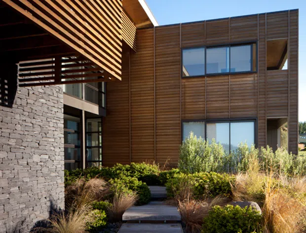 A contemporary multi-level building facade viewed from an outdoor approach. The structure combines warm horizontal timber cladding and deep timber screening with dark stacked-stone walling, large glazed windows, and projecting roof forms. A landscaped entry path with timber steps, gravel, grasses, shrubs, and low stone edging leads toward the building. Clear blue sky and strong sunlight emphasize the clean architectural lines and natural materials.