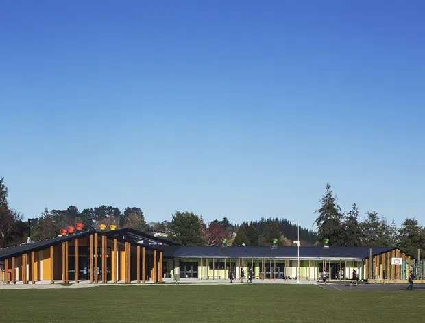 Wide exterior view of a low-rise contemporary educational or community building bordering a large grass field. The complex has a long, single-storey form with dark pitched roof planes, extensive glazing, warm timber vertical cladding and deep timber columns, with a connected lighter wing and covered outdoor circulation. Trees and a clear blue sky form the background.