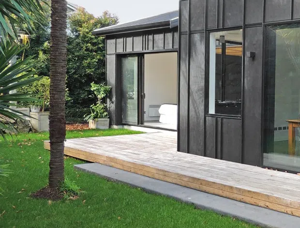 Exterior view of a contemporary single-storey extension or pavilion with dark vertical cladding, extensive black-framed glazing, and a raised timber deck opening onto a manicured lawn. Tropical-style palm planting frames the building, while a bright interior room is visible through the glazed doors.