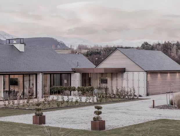A low, modern residential property viewed from the landscaped driveway, with multiple grey pitched roofs, pale weatherboard-style cladding, extensive floor-to-ceiling glazing, and an attached garage. The house sits in a spacious landscaped setting with clipped potted trees, ornamental grasses, gravel paving, and mountains visible in the distance under an overcast sky.