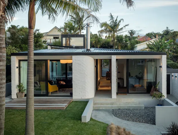 Front exterior view of a low-rise contemporary home in a tropical setting. The house has a flat, corrugated metal roof, white rendered and white painted brick walls, extensive floor-to-ceiling glazing, a rounded projecting facade element, and a small landscaped front garden with a gravel driveway. Palm trees and neighbouring homes surround the property.