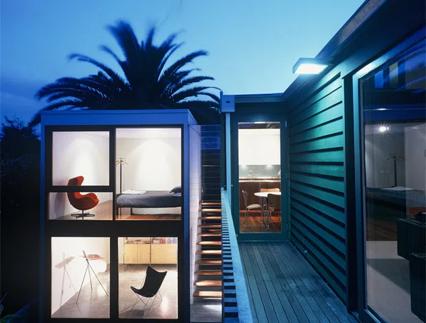 Twilight exterior view of a contemporary residential extension with dark horizontal cladding and timber decking. A narrow illuminated stair and walkway connect the main structure to a compact glazed pavilion, through which a bedroom and living area with mid-century furnishings are visible. A large palm tree silhouettes against the blue evening sky.
