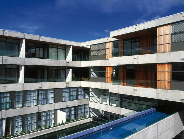 Exterior view of a multi-storey modern concrete building arranged around an internal courtyard. The facade features exposed concrete horizontal bands, extensive glazing, dark window frames, and warm vertical timber cladding panels. A narrow blue swimming pool or water feature is visible at the lower courtyard level.