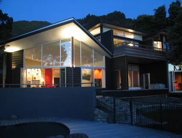 Twilight exterior view of a multi-level contemporary home with angular pitched roof forms, extensive floor-to-ceiling glazing, dark horizontal cladding, and a projecting upper-level balcony. A timber deck, pool edge, stone retaining elements, and landscaped hillside setting are visible.