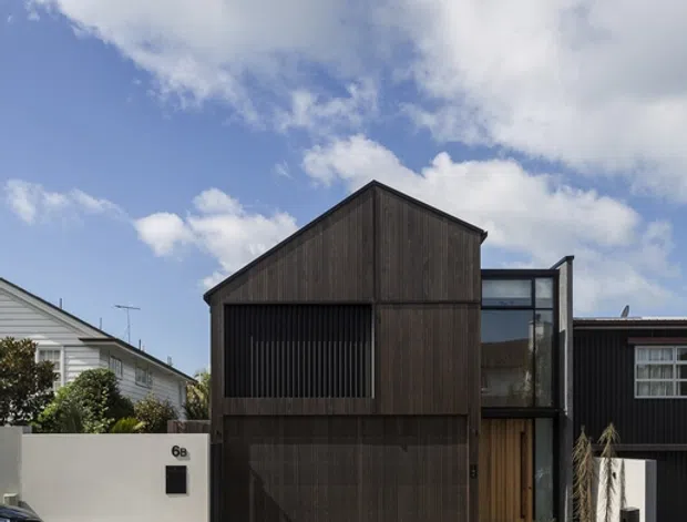 Street-facing photograph of a narrow contemporary two-storey residence with a steep gabled form, dark vertical timber cladding, a large integrated garage, upper-level screened glazing, and a glazed entry link. The home sits tightly between neighbouring houses along a paved suburban street under a bright partly cloudy sky.