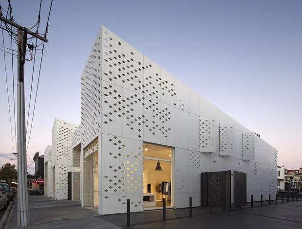 Street-level exterior view of a contemporary low-rise commercial building with a dramatic angular white facade. The envelope is composed of pale panel cladding punctured by dense geometric perforations, with large glazed shopfront openings and warm interior lighting. Utility poles and an urban streetscape frame the building.