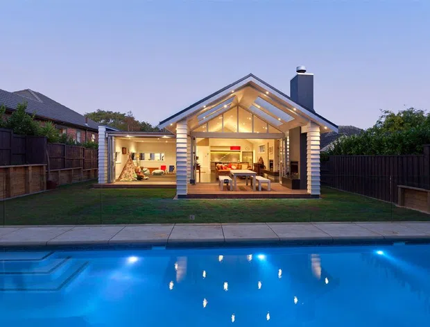 A contemporary single-storey backyard pavilion or extension photographed at dusk, opening directly onto a swimming pool. The structure has a prominent pitched gable roof with exposed timber rafters, a glazed triangular gable, warm recessed lighting, pale horizontal cladding, and a dark chimney. Through the wide opening, a dining area and adjoining lounge or activity space are visible. A paved pool edge, lawn, timber fencing, and neighbouring suburban homes surround the building.