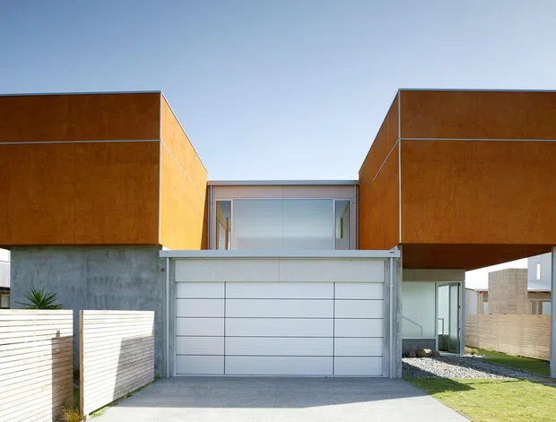 Street-facing exterior view of a contemporary multi-level residence composed of two projecting box-like upper volumes clad in warm rust-brown panels, set above a light grey garage and entry structure. Horizontal timber privacy fencing, neighbouring homes, a concrete driveway, and restrained landscaping are visible. The design features strong geometric forms, flat roofs, and a recessed glazed connection between the upper volumes.