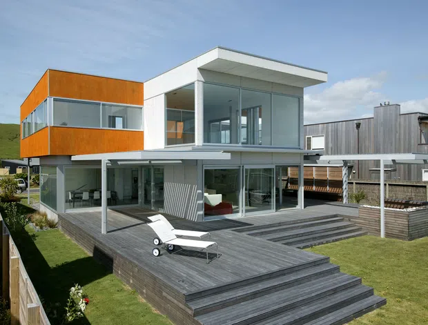 Exterior view of a contemporary multi-level residence with bold orange and white box-like volumes, extensive floor-to-ceiling glazing, and a broad grey timber deck with tiered steps. The house sits beside a sloping grassy site and includes a covered outdoor area and landscaped garden edges.