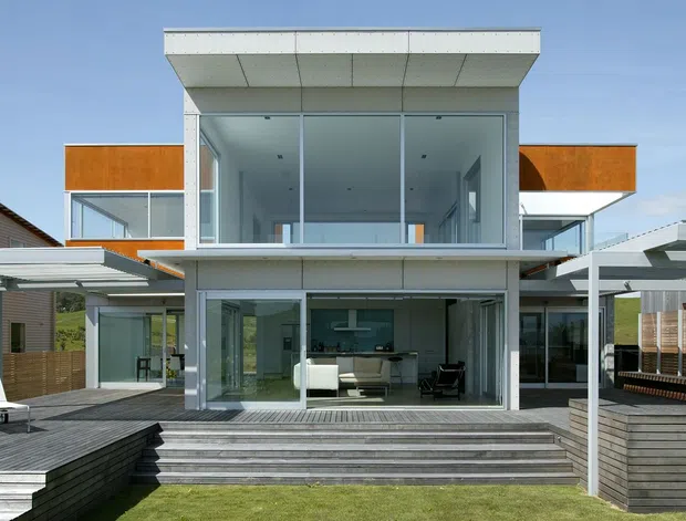 Exterior view of a contemporary multi-level residence with a strong central glazed volume, flat roof, projecting upper-floor box, and extensive floor-to-ceiling glazing. The house opens onto broad stepped decking and a landscaped backyard, with covered outdoor structures and neighbouring residences visible on either side.
