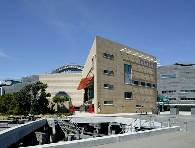 Exterior view of a large contemporary institutional or commercial complex with multiple connected buildings, angular and curved roof forms, extensive glazing, and exposed concrete plaza structures. The scene includes broad paved open space, stairways, ramps, and landscaped edges under a clear blue sky.
