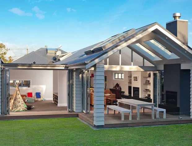 Exterior view of a contemporary residential home with a prominent glazed gable extension and large open folding doors. The extension contains a living area with a fireplace, shelving and dining table, while the adjoining room appears to be a children's playroom. The building features grey horizontal cladding, extensive glazing, skylights and a pitched roof, opening onto a lawn and timber deck.