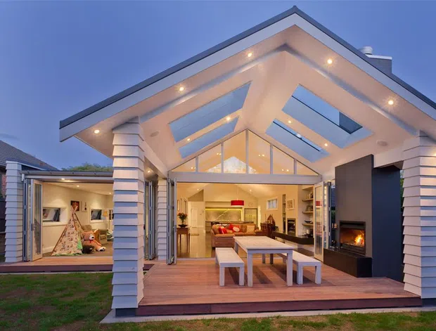 Exterior view of a contemporary residential extension at dusk, featuring a large open entertaining area with timber decking, broad glazed folding doors, a dramatic pitched roof, multiple skylights, and white horizontal-clad structural columns. The interior living room and a children's playroom are visible through the open glazing. A dark built-in fireplace wall and outdoor dining table define the covered terrace.
