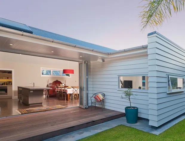 Single-storey residential courtyard-facing extension with pale horizontal weatherboard cladding, a blue low-pitched roof, and large folding glazed doors opening onto a timber deck. The open interior reveals a compact kitchen with island, dining area, and warm timber-toned flooring and ceiling. A small courtyard lawn, potted palm, and outdoor chair frame the foreground.