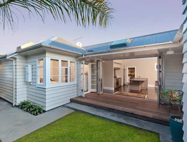 Exterior view of a light weatherboard home opening onto a timber deck and small lawn courtyard. A large glazed folding-door opening connects the deck to a bright kitchen and living area, with a blue-grey metal hipped roof and coastal-style detailing.