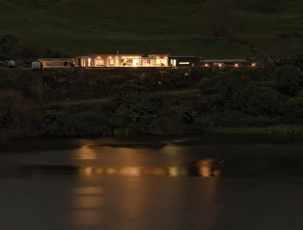 Night-time exterior view of a long, low modern building illuminated warmly along a dark, vegetated hillside beside calm water. The building’s lights create elongated golden reflections across the water, while the surrounding landscape remains shadowed and largely natural.