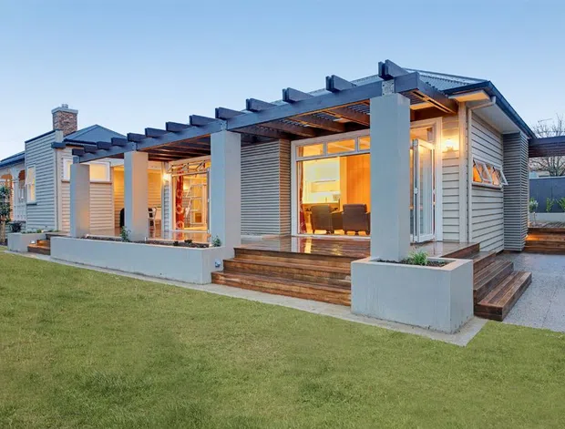 Exterior view of a contemporary single-storey home at dusk, featuring a low-pitched roof, horizontal light-grey cladding, broad glazed openings, timber decking and steps, and a deep timber pergola with exposed beams. Warm interior lighting is visible through the windows and open living area, with a neatly landscaped lawn in the foreground.