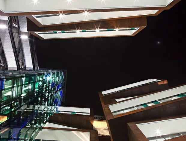 Upward-looking architectural view of a modern multi-storey building or commercial complex at night. Dark glazed façades, reflective glass volumes, projecting horizontal roof or canopy elements, and illuminated recessed ceiling panels create a dramatic composition. The image focuses on exterior architecture rather than an interior room.
