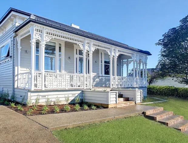 Exterior view of a restored single-storey timber villa or bungalow with ornate white fretwork, decorative veranda posts and balustrades, weatherboard cladding, a corrugated metal roof, and large sash-style windows. The home sits within a neat suburban garden with concrete paths and brick steps.