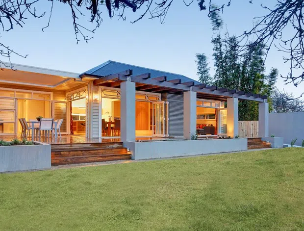 Single-storey contemporary residential house viewed from the garden, with extensive warm-lit glazing, timber-clad walls, a broad timber deck, concrete garden planters, and a pergola-like covered outdoor structure supported by square concrete columns. The building has a low-pitched metal roof and a strong indoor-outdoor connection opening onto a lawn.