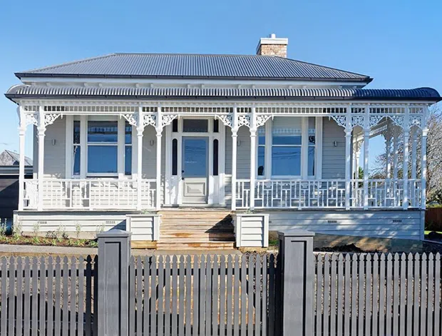 Street-facing exterior view of a single-storey heritage-style house with an ornate white timber veranda, decorative fretwork, turned posts, balustrades, weatherboard cladding, and a corrugated hipped roof. The facade includes a central entry door, symmetrical windows, front steps, and a contemporary dark picket fence.
