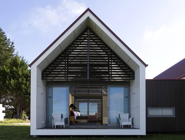 Front exterior view of a modern gable-roof house with a white rendered or panelled facade, deep covered entry, extensive glazing, and a prominent dark timber louvre screen filling the triangular gable. A darker ribbed-cladding wing connects to the right, with landscaped grounds and a neighboring white building partially visible.