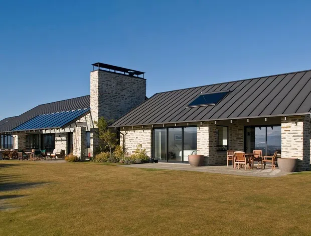 Single-storey contemporary stone-clad residence with multiple pitched metal-roof volumes, a prominent central chimney, skylight, extensive glazing, and a broad landscaped lawn. Outdoor dining and covered verandah areas connect the house to the garden.