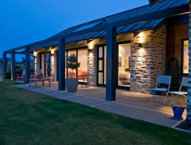 A single-storey modern residence photographed at dusk, with a long covered outdoor verandah supported by dark steel posts and beams. The facade combines extensive glazing and doors with irregular horizontally coursed natural stone walls, warm recessed lighting, and a dark standing-seam metal roof. Outdoor seating, planters, and a lawn occupy the foreground, while softly lit interior rooms are visible through the glazing.