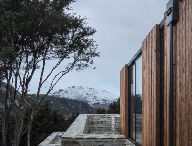 Exterior view of a contemporary mountain home with vertically clad timber walls, expansive dark-framed glazing, and a stepped natural-stone terrace or retaining wall. Snow-covered mountains and native vegetation form the background, emphasizing the building's elevated alpine setting.