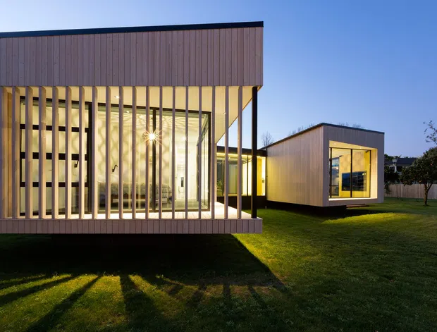 Exterior dusk view of a contemporary single-storey modular-style home composed of rectilinear volumes. The building features vertical timber cladding, extensive floor-to-ceiling glazing, and a timber slatted screen forming a sheltered outdoor edge. Warm interior lighting contrasts with the cool evening light, while the separate projecting volume and open lawn emphasise the pavilion-like composition.