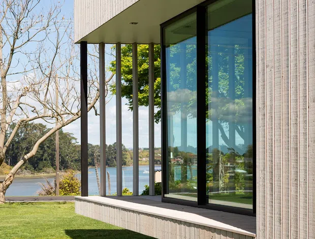 Exterior architectural photograph of a contemporary rectilinear building with vertically textured light cladding, a deep cantilevered overhang, slender dark structural columns, and large floor-to-ceiling glazed sliding doors. The building sits above a manicured lawn beside a calm waterfront, with trees and distant shore visible through and around the glazing.