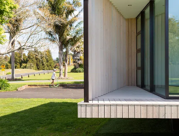 Exterior architectural view of a contemporary building with a projecting covered terrace or veranda. The facade is clad in pale vertical timber boards, with dark structural posts, recessed downlights, and extensive dark-framed floor-to-ceiling glazing. The building is set within a sunny landscaped suburban setting with lawn, mature trees, and a nearby street.