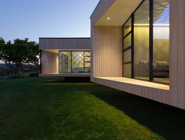 Exterior dusk view of a contemporary, single-storey building composed of offset rectangular volumes. The facade features vertical light timber cladding, extensive floor-to-ceiling black-framed glazing, projecting cantilevered sections, and warm interior lighting. The building sits within a broad lawn and landscaped setting.